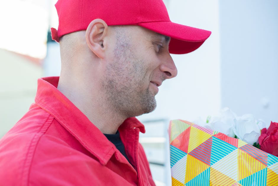 A delivery person wearing a red cap and matching red jacket is holding a colorful gift bag filled with white and red flowers. The flowers are fresh and vibrant, with delicate petals and green stems vi