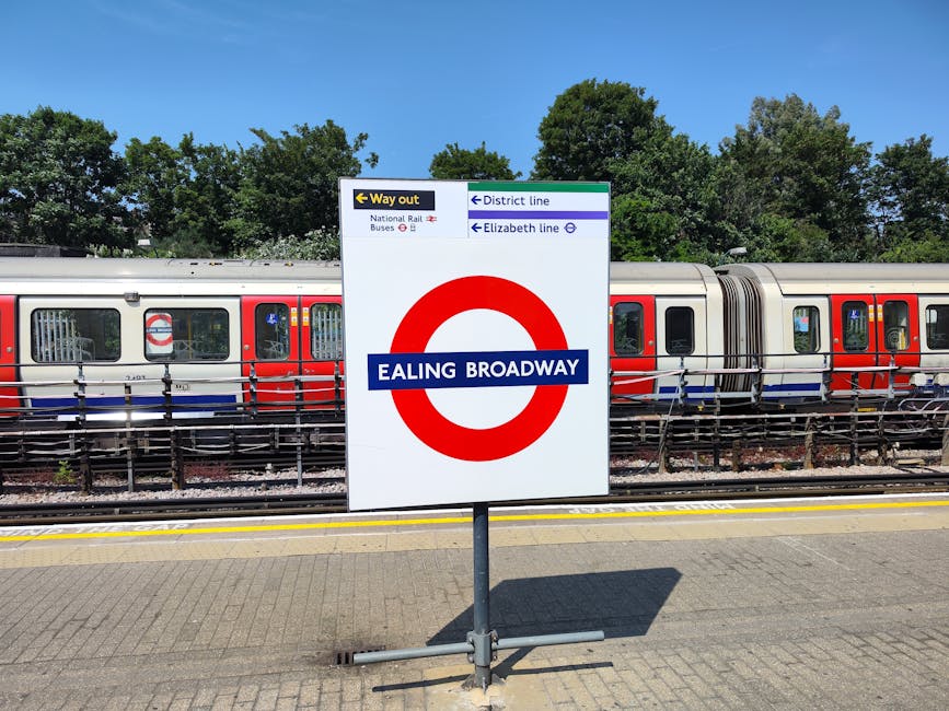 A close-up view of the Ealing Broadway underground station sign with a London Underground train in the background. The sign displays the station name in white text on a blue background, encircled by a