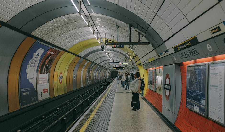 A group of passengers waiting on the platform at Green Park station, an underground tube station with a curved ceiling featuring yellow, gray, and white panels. The platform has a yellow safety line a