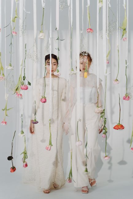 Two women dressed in elegant, light-colored lace dresses stand behind a hanging installation of pink, red, and purple flowers suspended from white strings. The flowers are arranged along green stems w