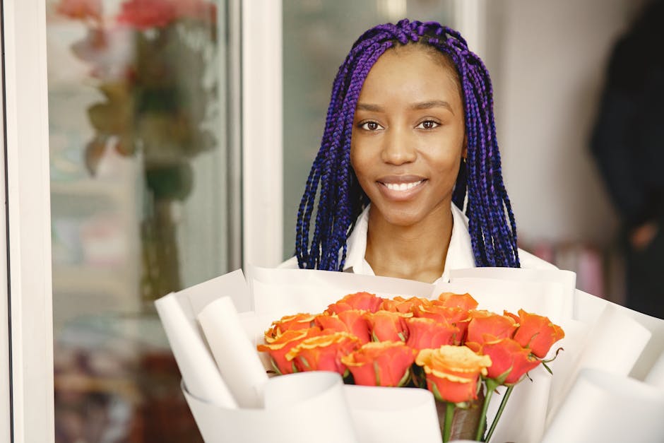 A young woman with dark skin and vibrant purple braided hair, smiling gently, holds a large bouquet of fresh, orange and pink roses wrapped in white paper. She stands indoors, possibly behind a floral