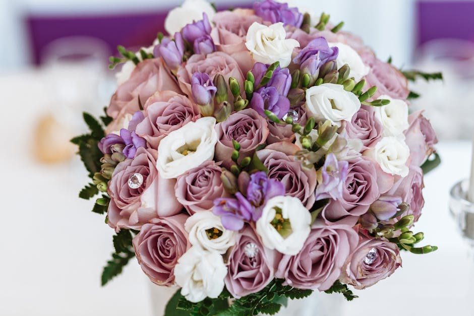 A close-up view of a floral bouquet arranged with soft pink and lavender roses, accented by white lisianthus and purple freesias, with green foliage peeking through. The flowers are fresh, with velvet