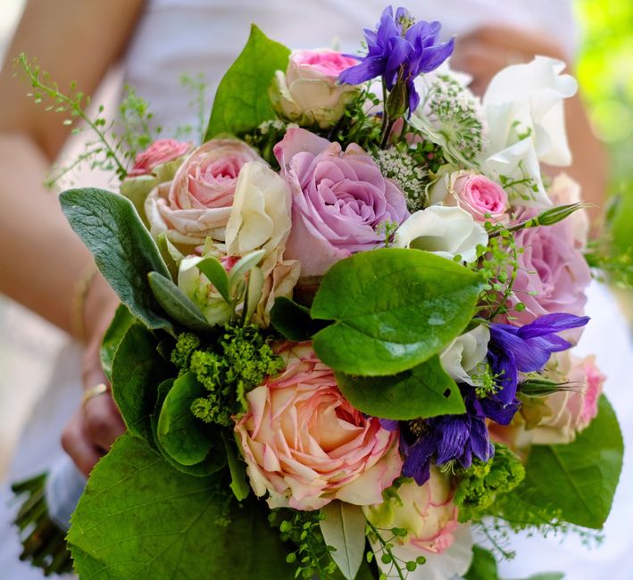 A close-up view of a floral bouquet featuring pink and cream roses, interspersed with purple irises and small white filler flowers, complemented by lush green leaves and stems. The arrangement appears
