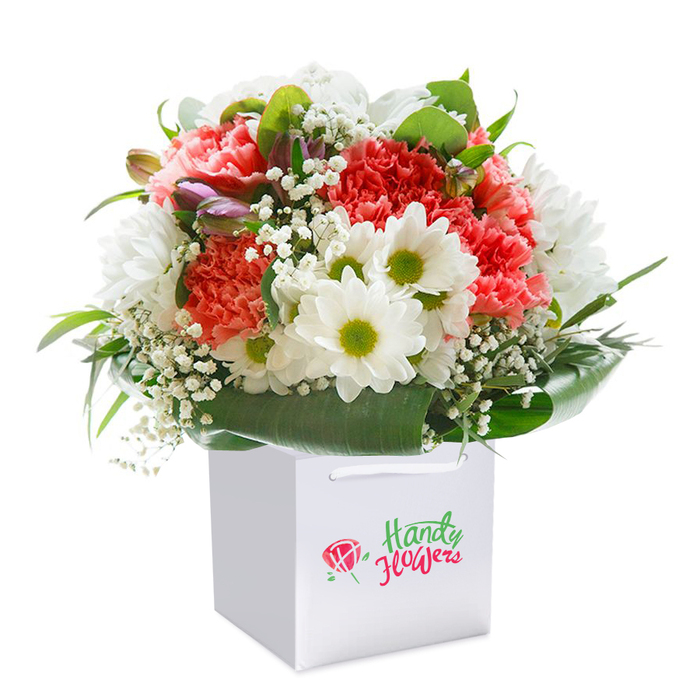 Arranged with care by the skilled hands of Woodside Park Florist, this visually striking bouquet sits upright in an elegant white square gift bag, untouched by distraction thanks to its seamless white background. The floral selection is unmistakably opulent: coral, peach, and deep red carnations-recognisable by their lavishly frilled petals-combine to create a domed centre bursting with cheerful energy, reminiscent of the vivid flowerbeds often seen in Woodside Park's communal gardens. Counterbalancing these, a scattering of bright daisies with gleaming green-yellow eyes brings a sprightly innocence to the piece, softened further by billows of wispy white gypsophila that distinguish the bouquet's delicate character. Subtle closed buds tinged with purple and rose hues add a quiet intrigue on the arrangement's left, hinting at life's unfolding surprises. The blooms nestle within a layer of broad, shining green leaves, their polished tips curving to hold the arrangement like a natural cradle-emphasising freshness, structure, and a touch of classic elegance. This rounded, full composition feels especially generous, reflecting the style and hospitable traditions of Woodside Park locals. The strong, matte-white ribbon handles arch above the bag, making the bouquet easy to deliver to neighbours, friends, or community celebrations. Lit to perfection, the flower colours and vivid greenery leap forward, expressing both the high standards and heartfelt touch that define everyday floristry in Woodside Park-a versatile gift for birthdays, anniversaries, or simply to share a little natural splendour, straight from your local florist.